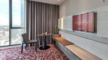 A hotel room with a large window, a TV mounted on the wall, a round table with a chair, and a bench.
