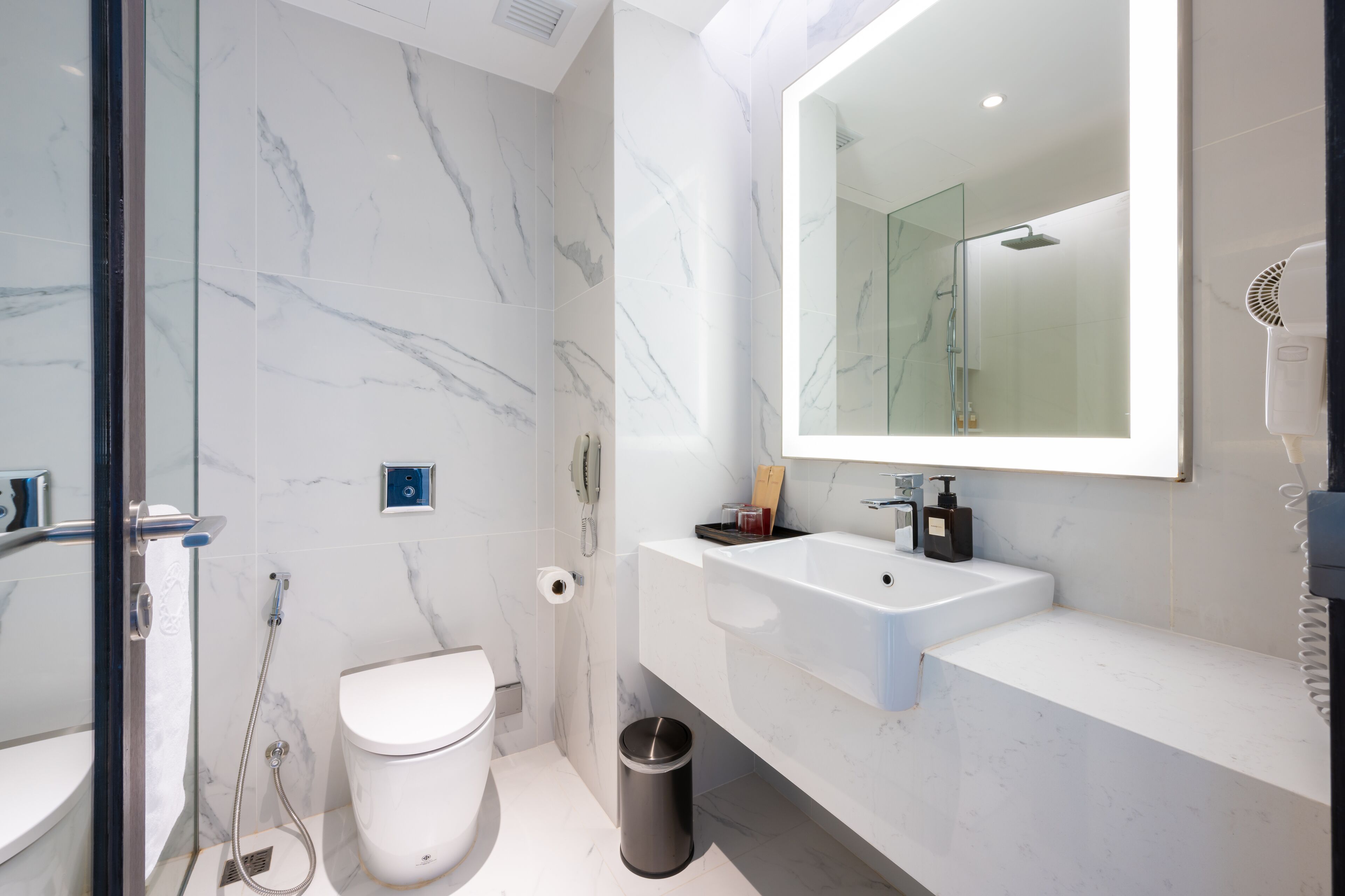 Un baño moderno con cabina de ducha de vidrio, inodoro blanco, lavamanos con espejo y cubo de basura.