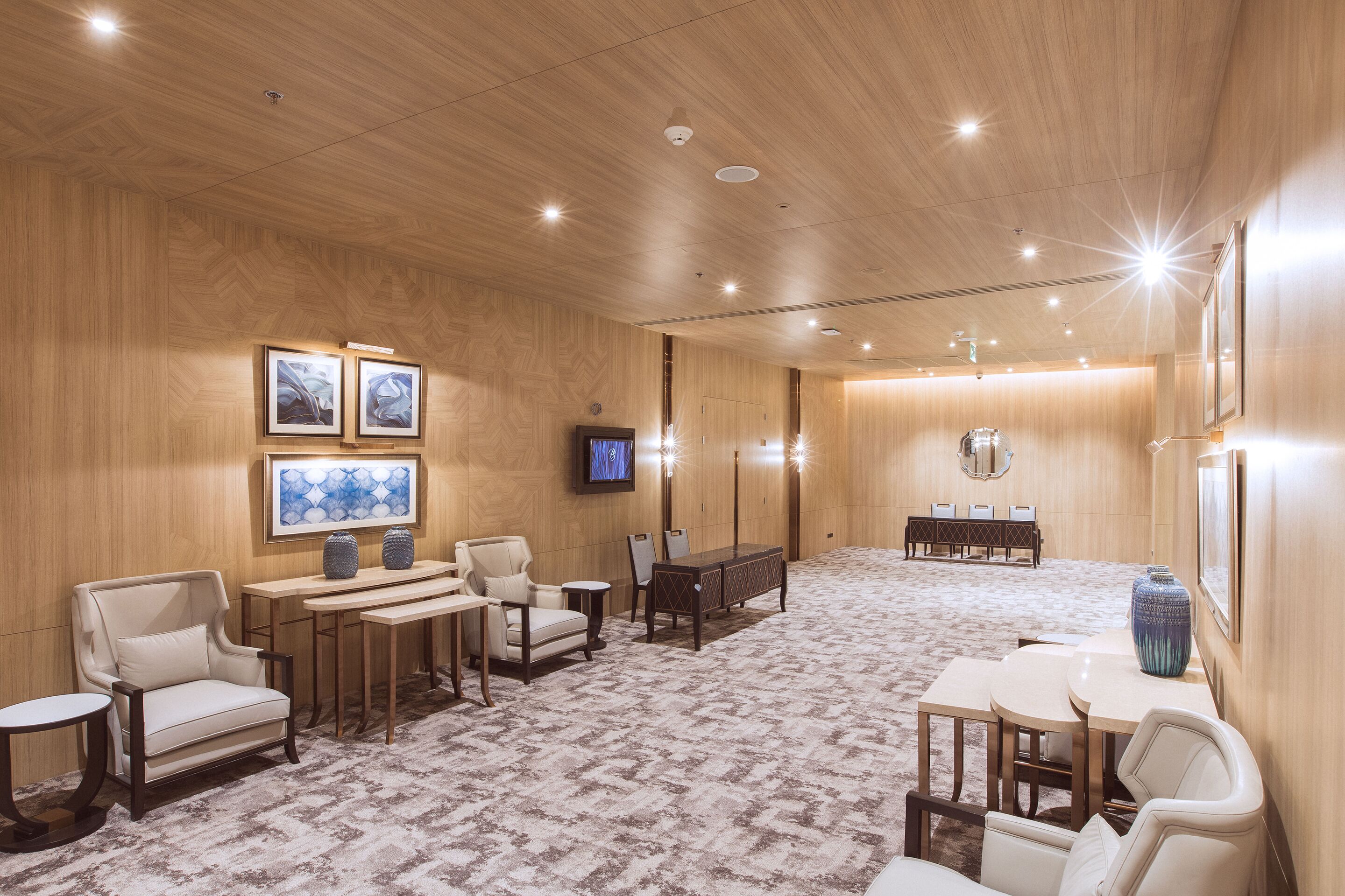 Una habitación amplia con paredes de paneles de madera, suelo alfombrado, televisor montado en la pared y un mostrador de recepción al fondo.
