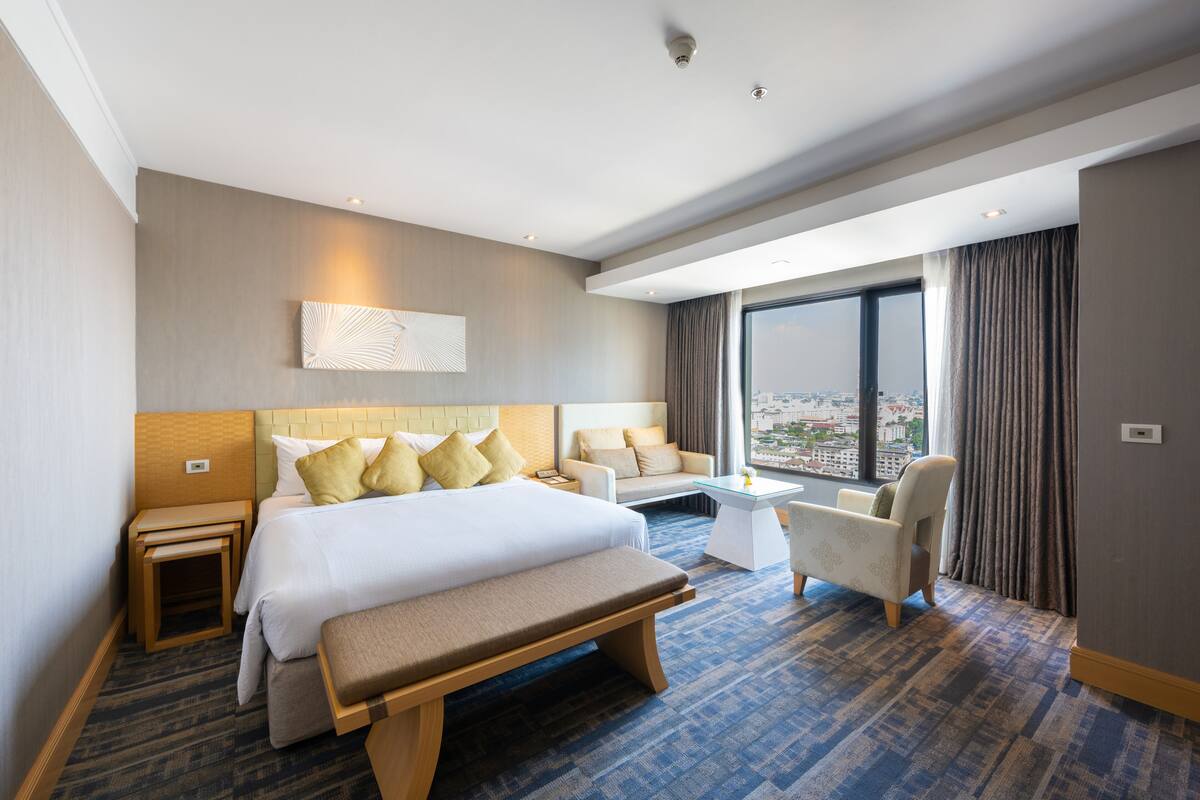 A hotel room with a large bed, a sofa, a bench, a small table, and a view of the city through the window.