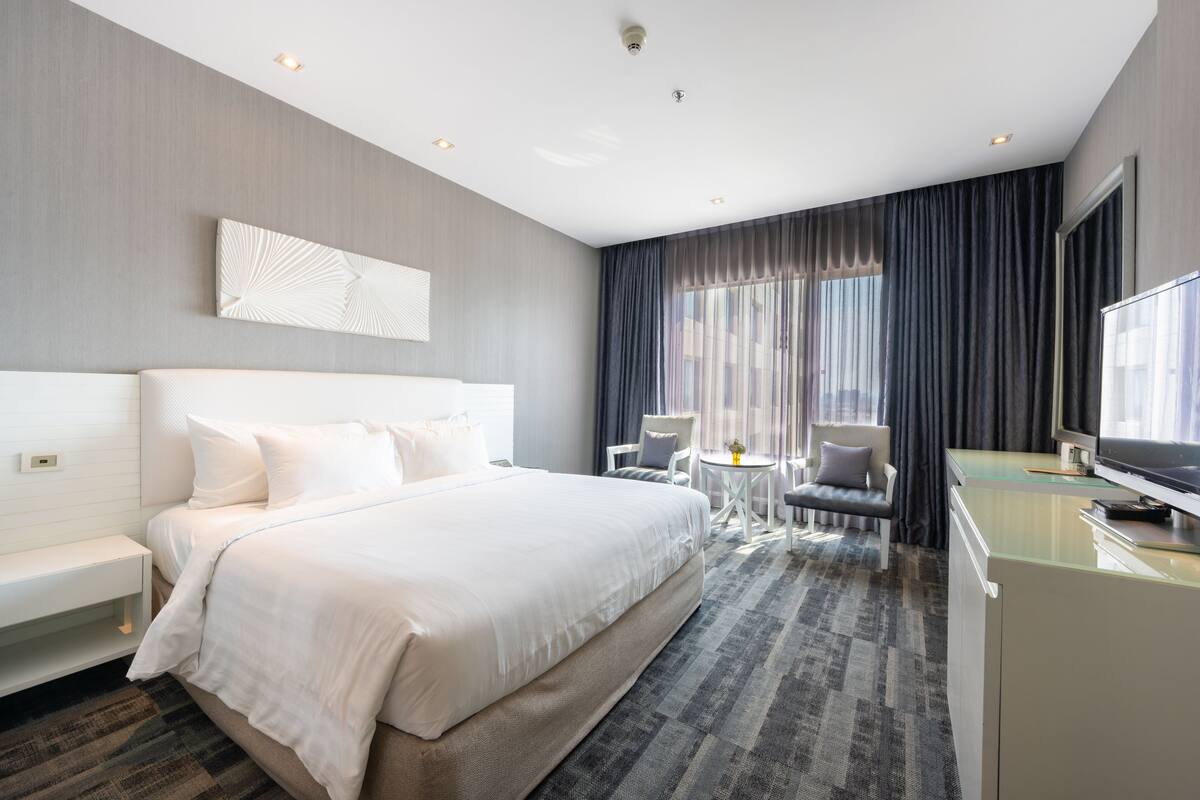 A modern hotel room with a large bed, a desk, a television, and a seating area with a view of the city.
