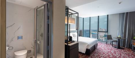 A hotel room with a bed, a desk, a chair, a bathroom with a toilet and shower, and a city view.