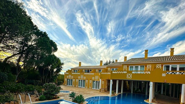 Una piscina con cubierta de piedra, un edificio con paredes decoradas y un cielo despejado con nubes.