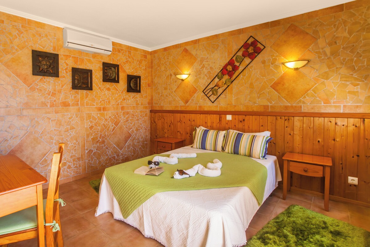A hotel room with a stone-patterned wall, a wooden headboard, a bed with a green blanket, a wooden nightstand, a green rug, and a wooden desk with a chair.