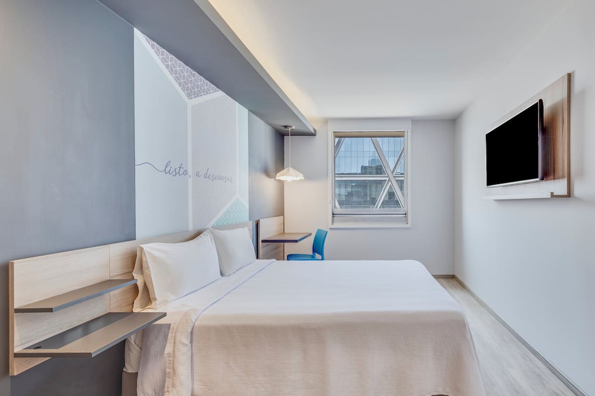 Ein modernes Hotelzimmer mit Bett, Schreibtisch, Stuhl, Fernseher und Fenster mit Stadtblick.