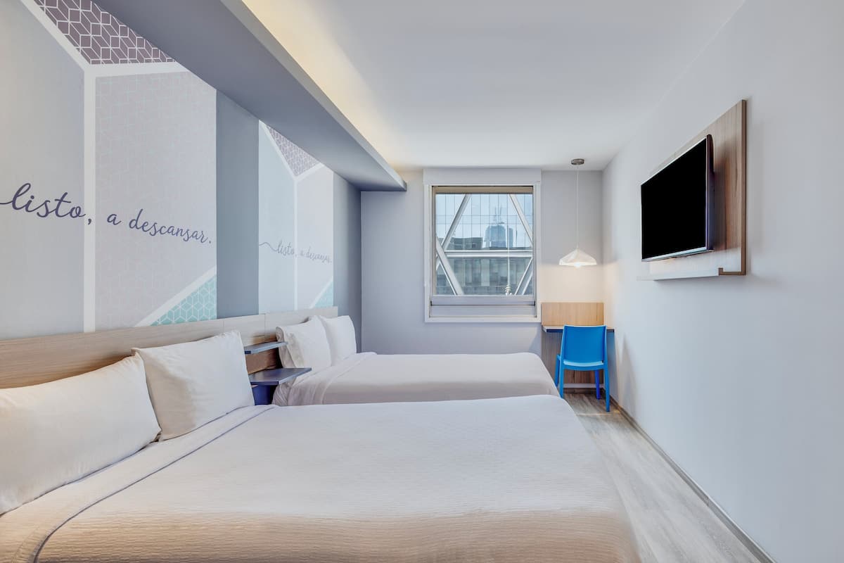 Ein Hotelzimmer mit zwei Betten, einem Fernseher, einem Fenster mit Blick auf die Stadt und einer Wand mit Text.