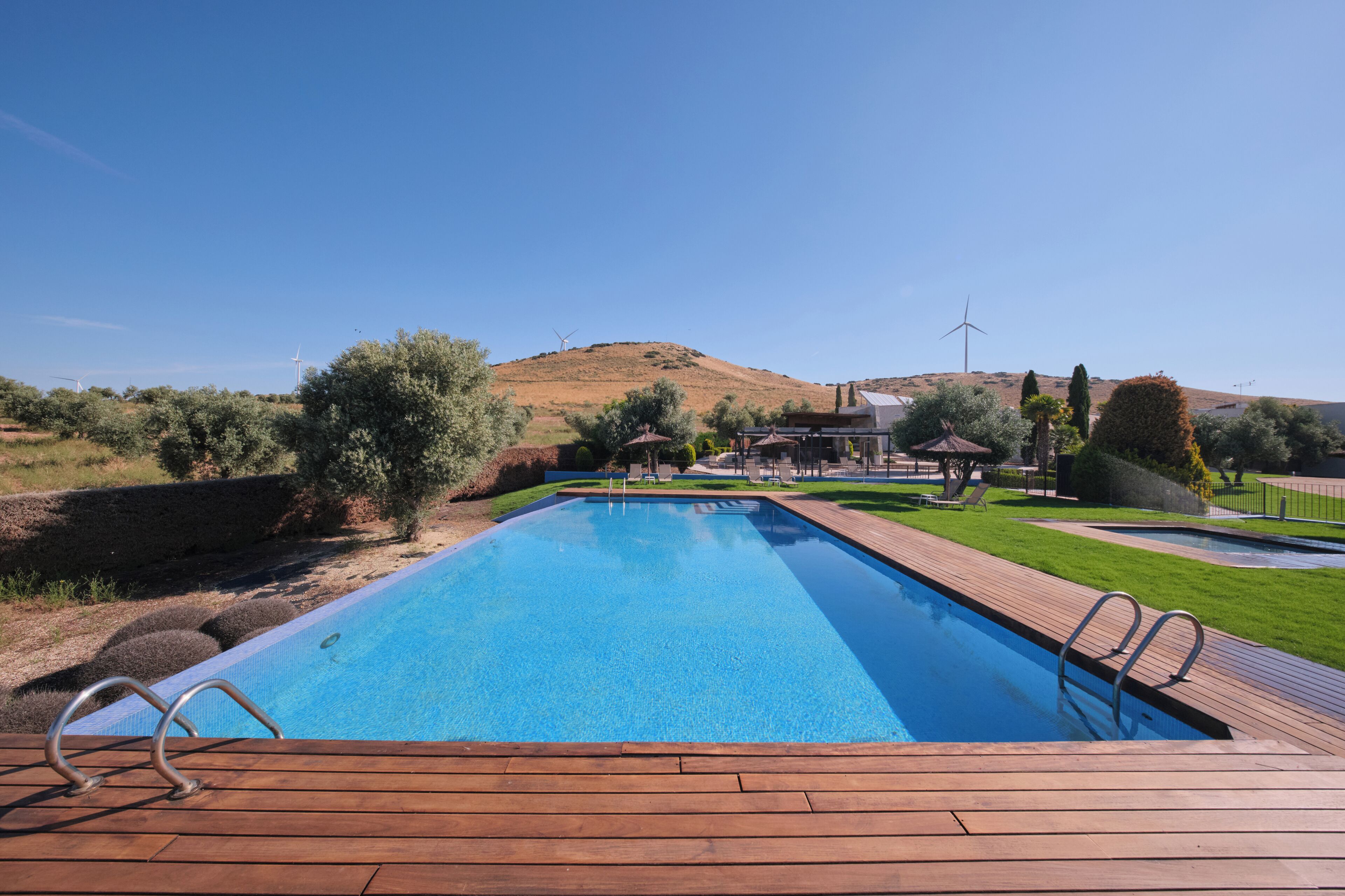Piscina retangular com um deck de madeira, cercada por vegetação e colinas.