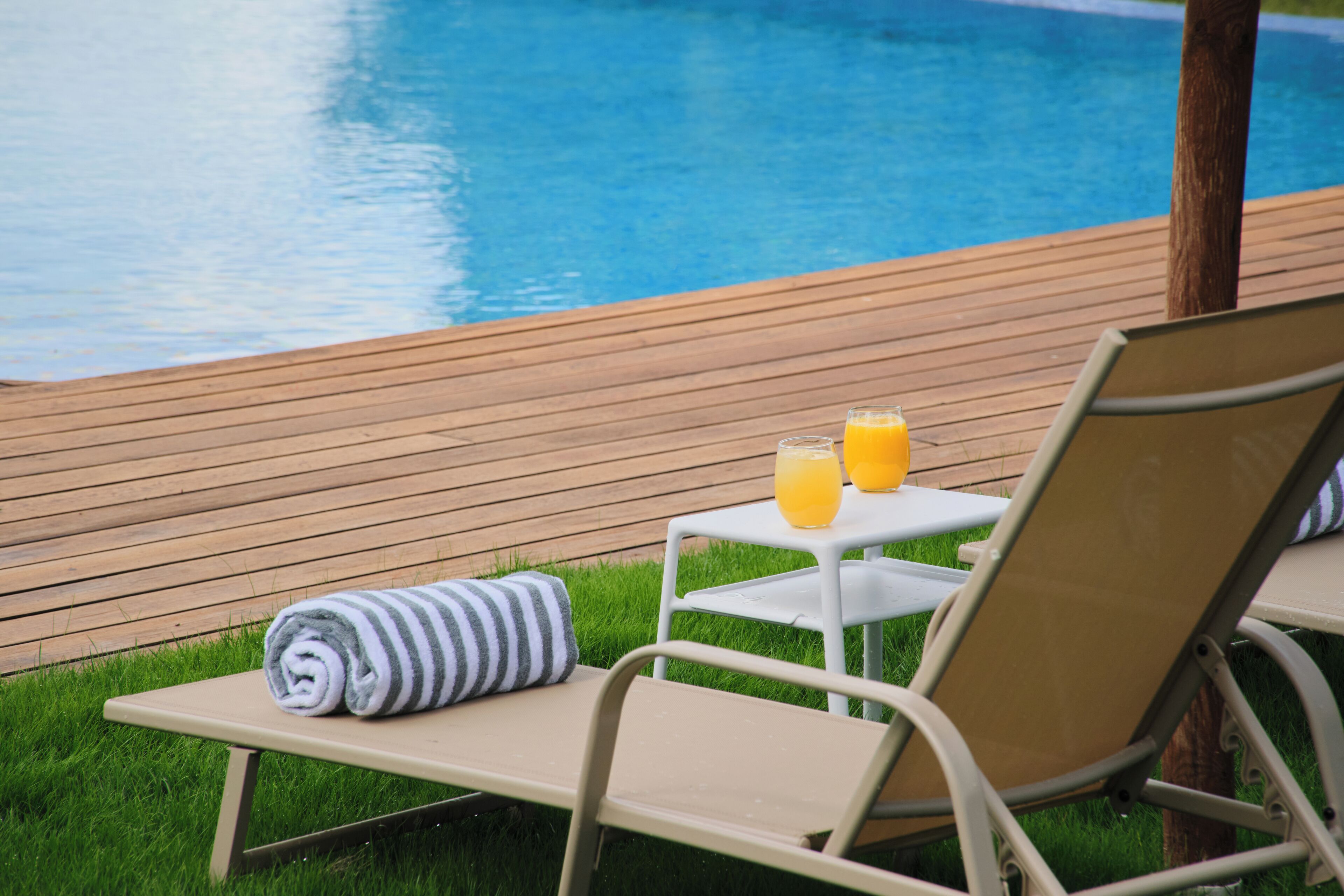 A poolside area with a wooden deck, a lounge chair, a small table with two glasses of orange juice, and a rolled towel on the deck.