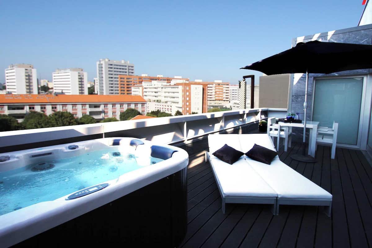 Een terras op het dak met een jacuzzi, lounge stoelen en een tafel met parasol.