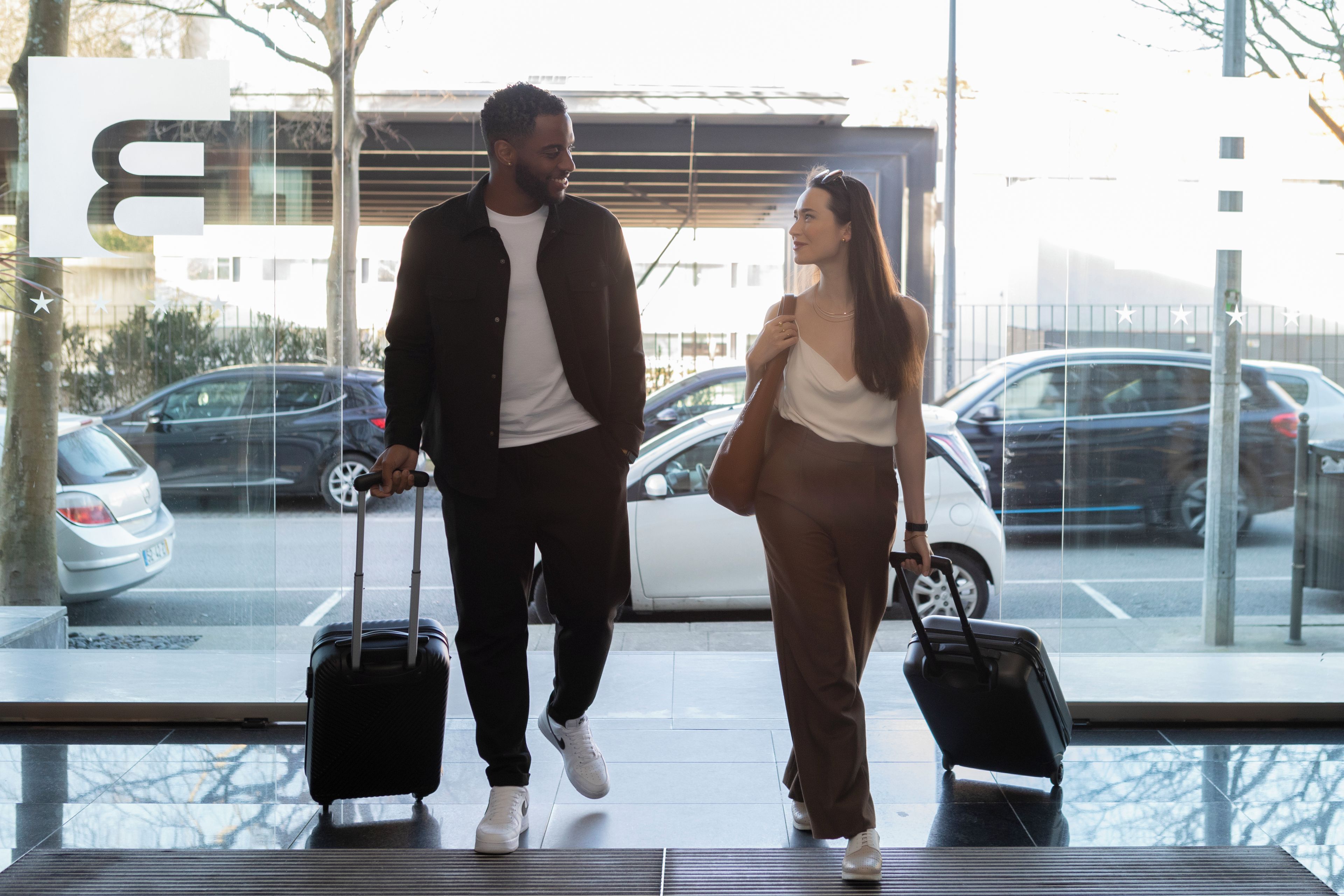 A man and a woman walking through a glass door with luggage, a parking lot with cars visible in the background.
