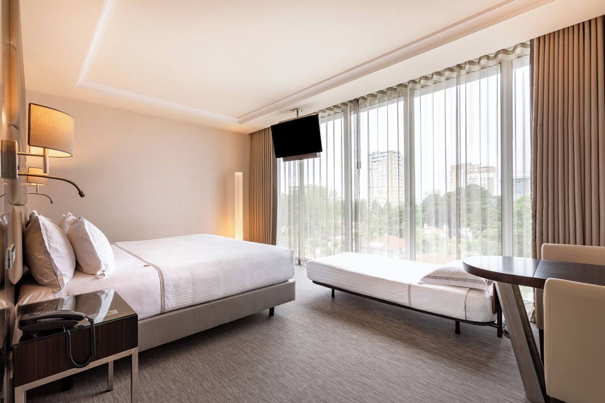 Een hotelkamer met een bed, nachtkastje, lamp, televisie en een raam met gordijnen.