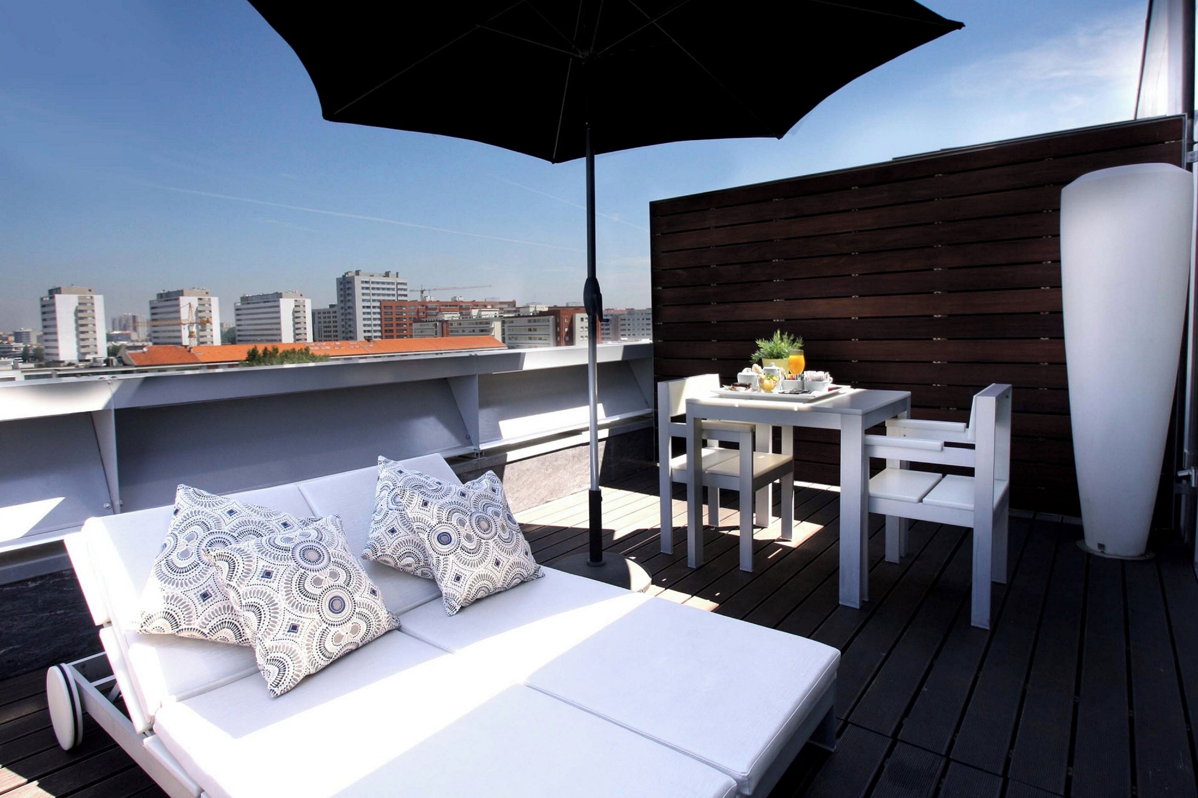 Een dakterras met een witte ligstoel, gestreepte kussens, een tafel met stoelen en een grote parasol.