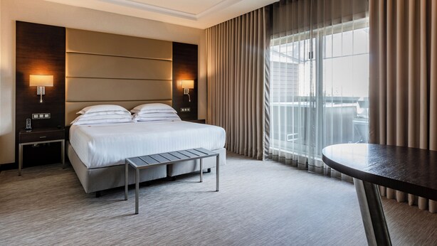 Ett modernt hotellrum med en stor säng, sängbord, ett litet bord och utsikt över staden genom stora fönster.