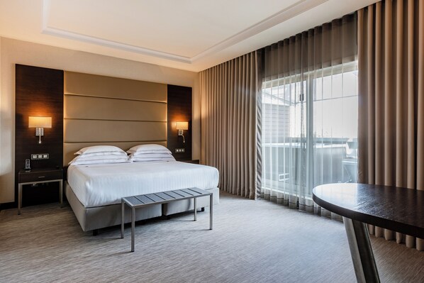 Habitación de hotel moderna con una cama grande, mesitas de noche, una pequeña mesa y vistas a la ciudad a través de ventanas amplias.
