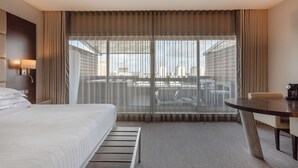 Ett hotellrum med en säng, ett skrivbord och utsikt över stadsbilden genom tunna gardiner.