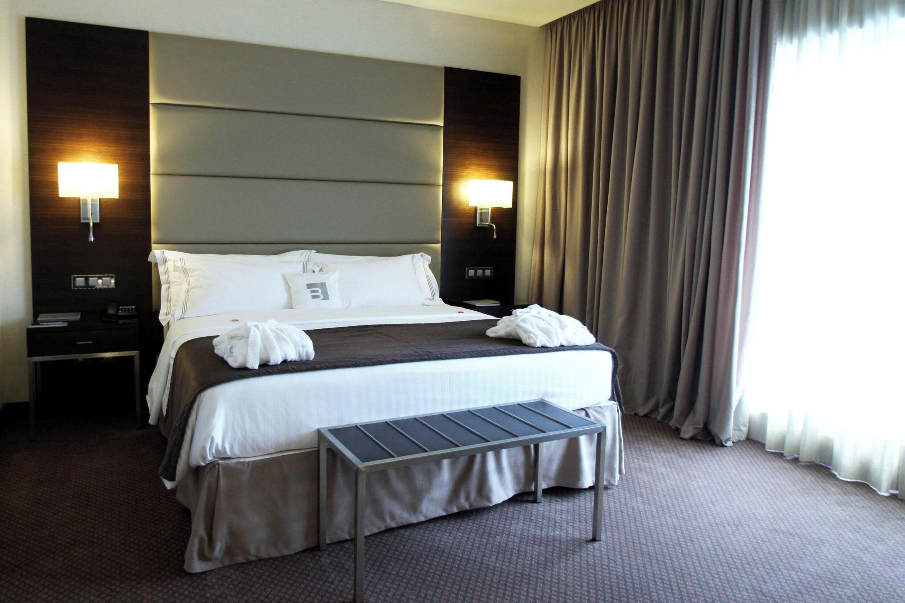 A hotel room with a large bed, two bedside tables, a desk, a chair, and a window with curtains.