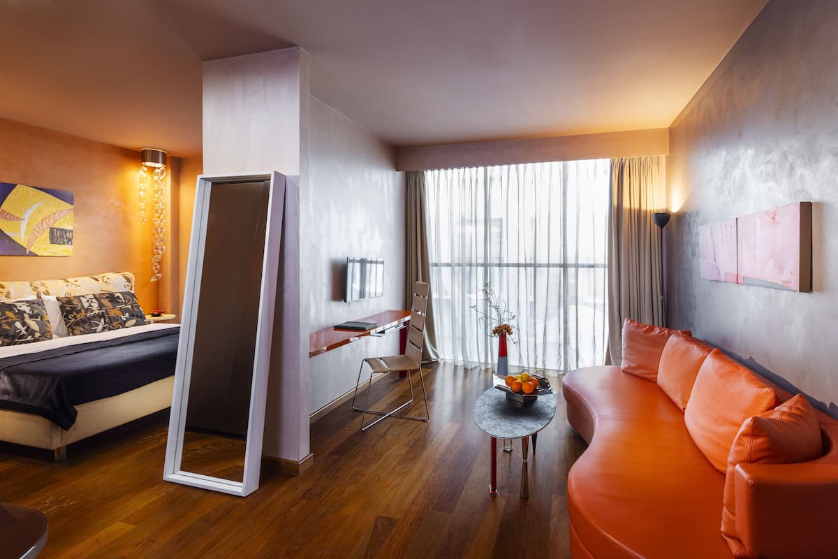 Una habitación de hotel moderna con cama, sofá, mesa de comedor y un gran ventanal con cortinas.