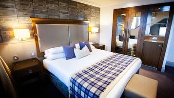 Een hotelkamer met een groot bed, een houten hoofdbord, een geruit sprei, een nachtkastje, een lamp en een kledingkast met spiegel.