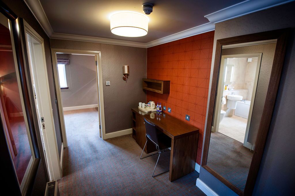 A hotel room with a wooden desk, a chair, a mirror, and a small shelf with toiletries.