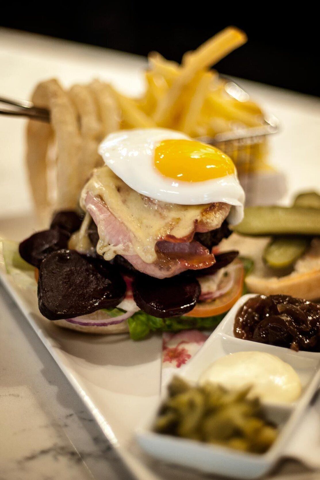 A burger with a fried egg on top, surrounded by pickles and a small bowl of sauce.