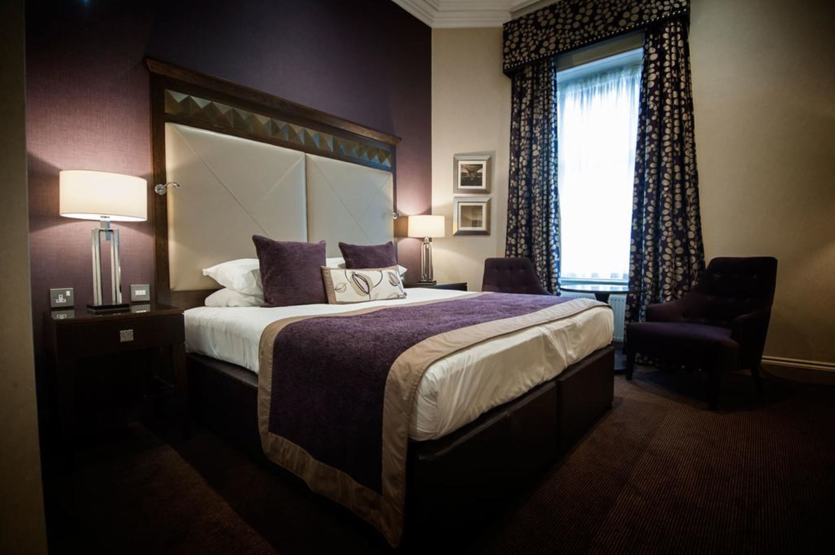 Quarto de hotel com uma cama grande, duas mesas de cabeceira, uma cadeira e uma janela com cortinas estampadas.