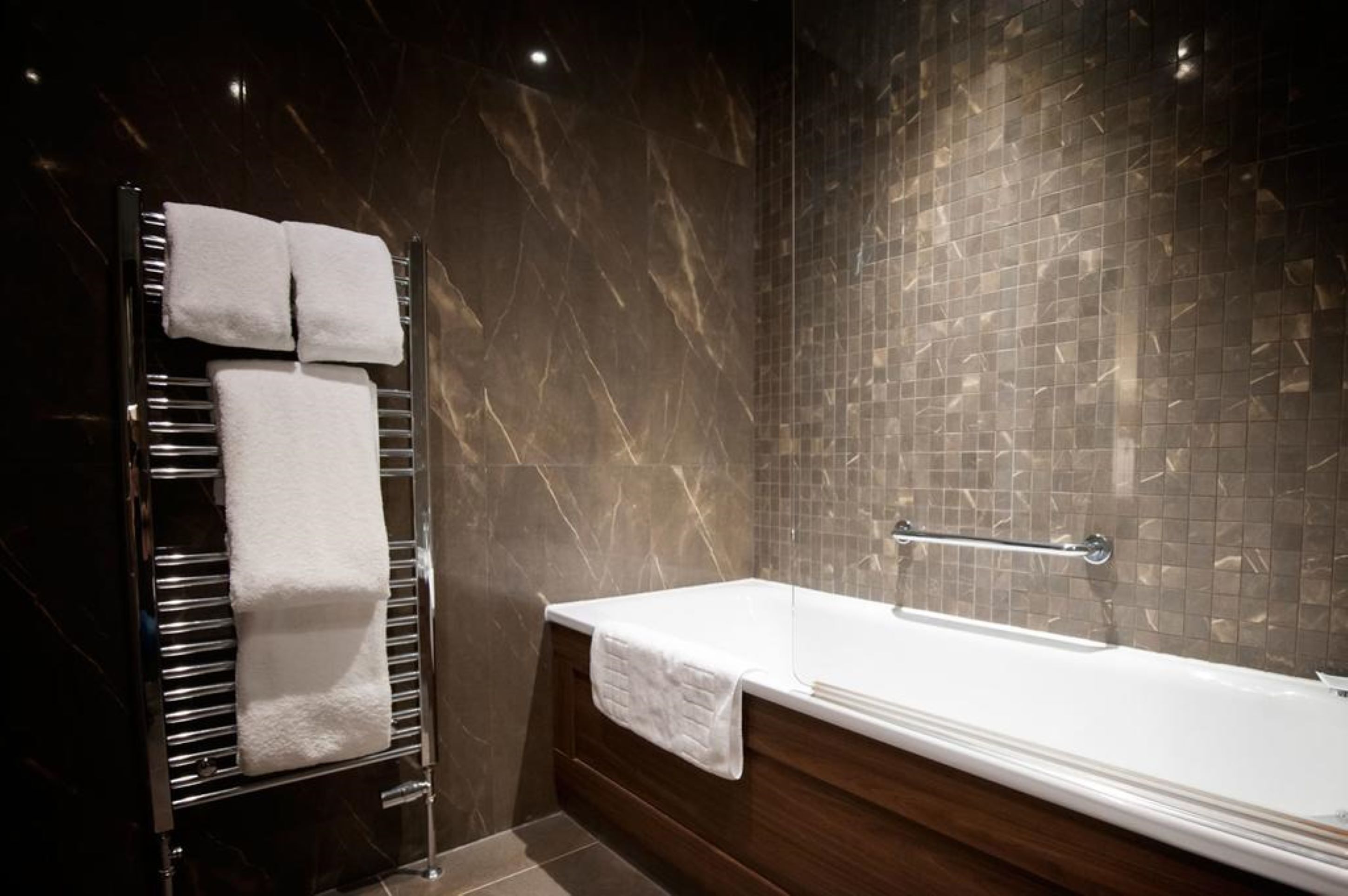A modern bathroom with a marble wall, a glass-enclosed bathtub, and a towel rack with white towels.