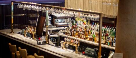 Zona de bar con mostrador de mármol, varios taburetes y una amplia variedad de botellas de licores, además de un espejo grande que refleja el bar.
