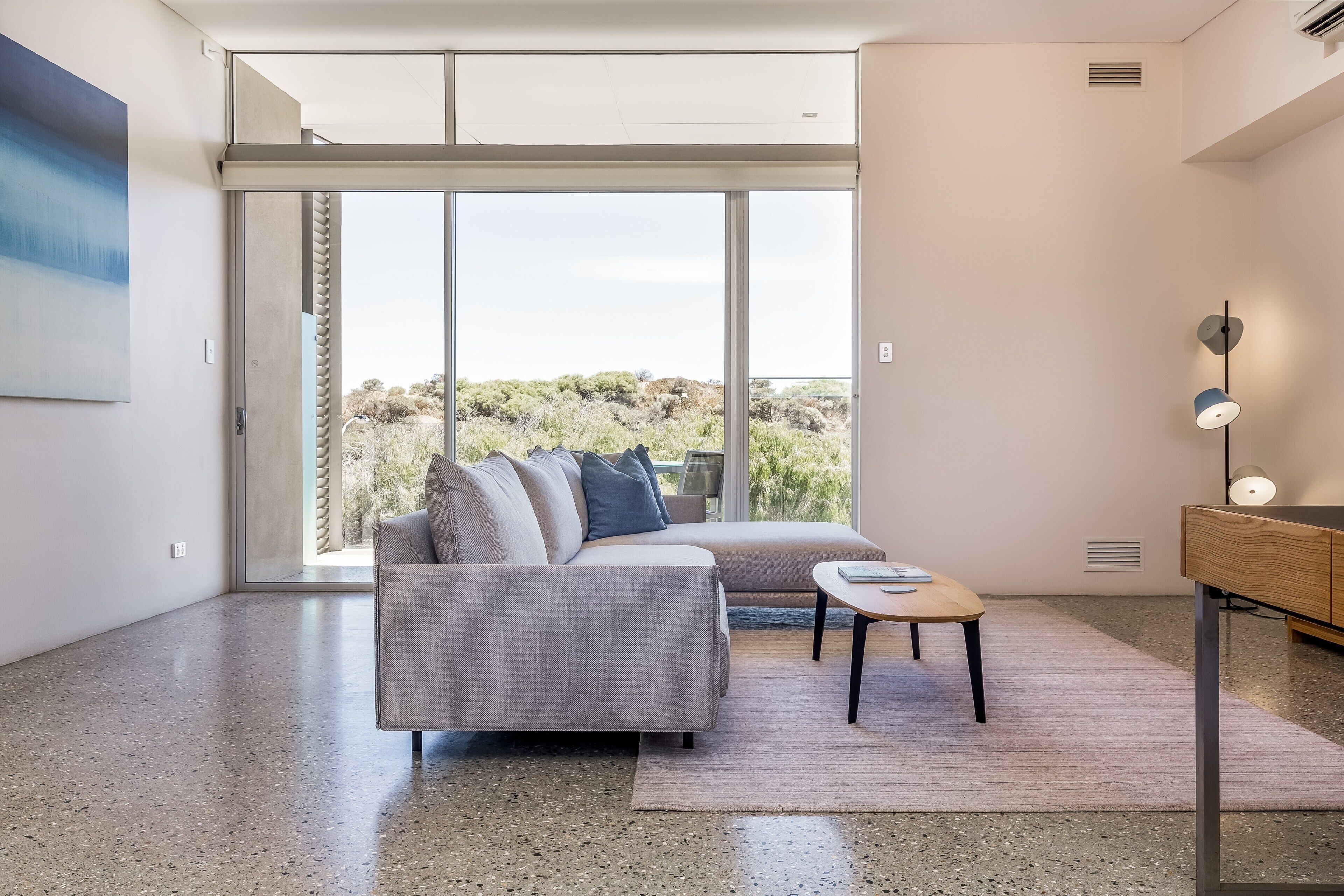 Un salon moderne avec un canapé gris, une petite table en bois et une grande porte-fenêtre coulissante donnant sur un balcon avec vue sur un paysage désertique.