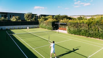 Tennisbane med to spillere, omgivet af grønt og en bygning i baggrunden.