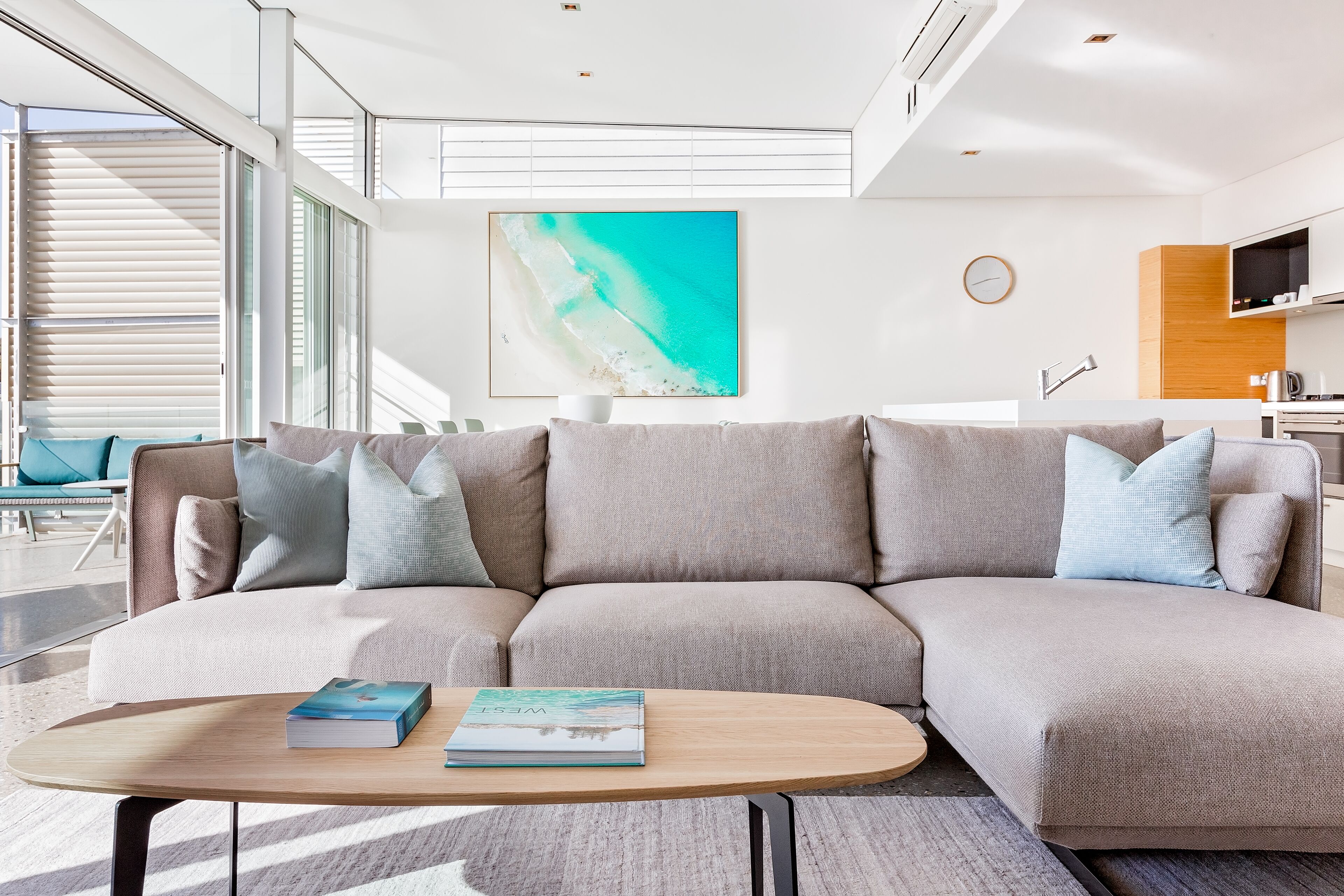 A modern living room with a beige sectional sofa, a wooden coffee table, and a large window with blinds.