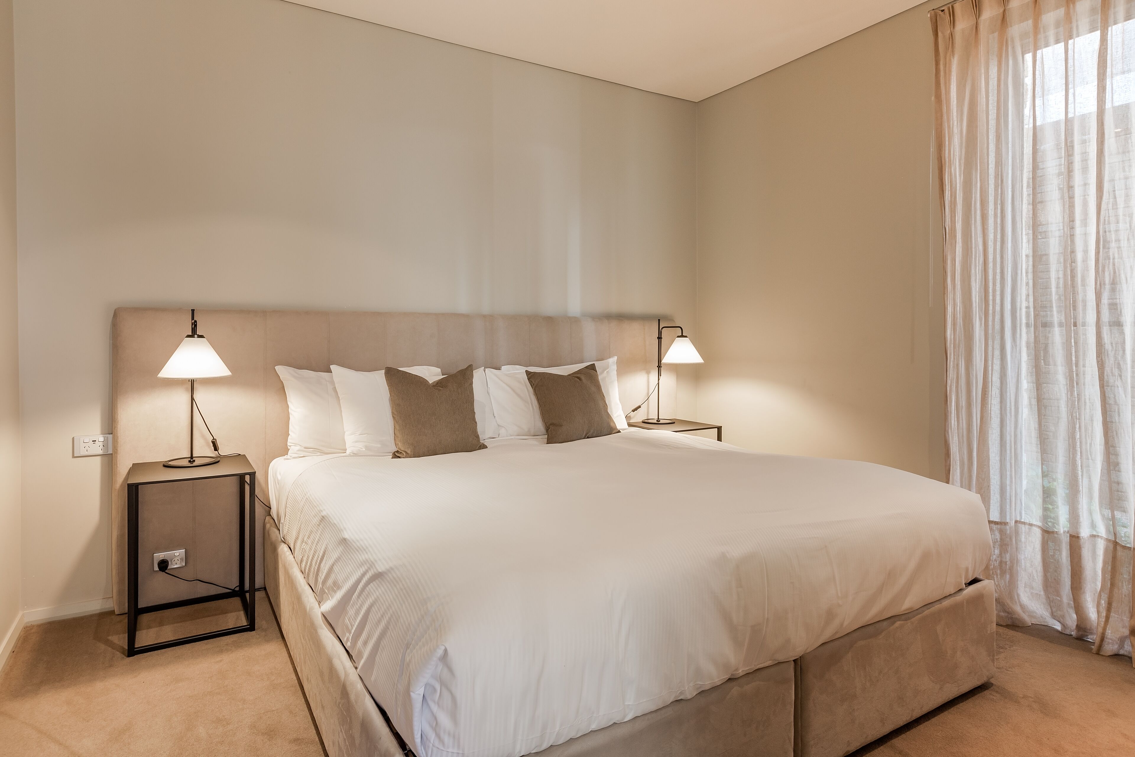A neatly made bed with a headboard, two bedside tables with lamps, and a window with sheer curtains.