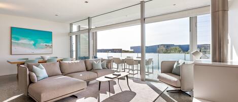 A modern living room with a large sofa, coffee table, and chairs, a dining area with a table and chairs, and large windows offering a view of the city.