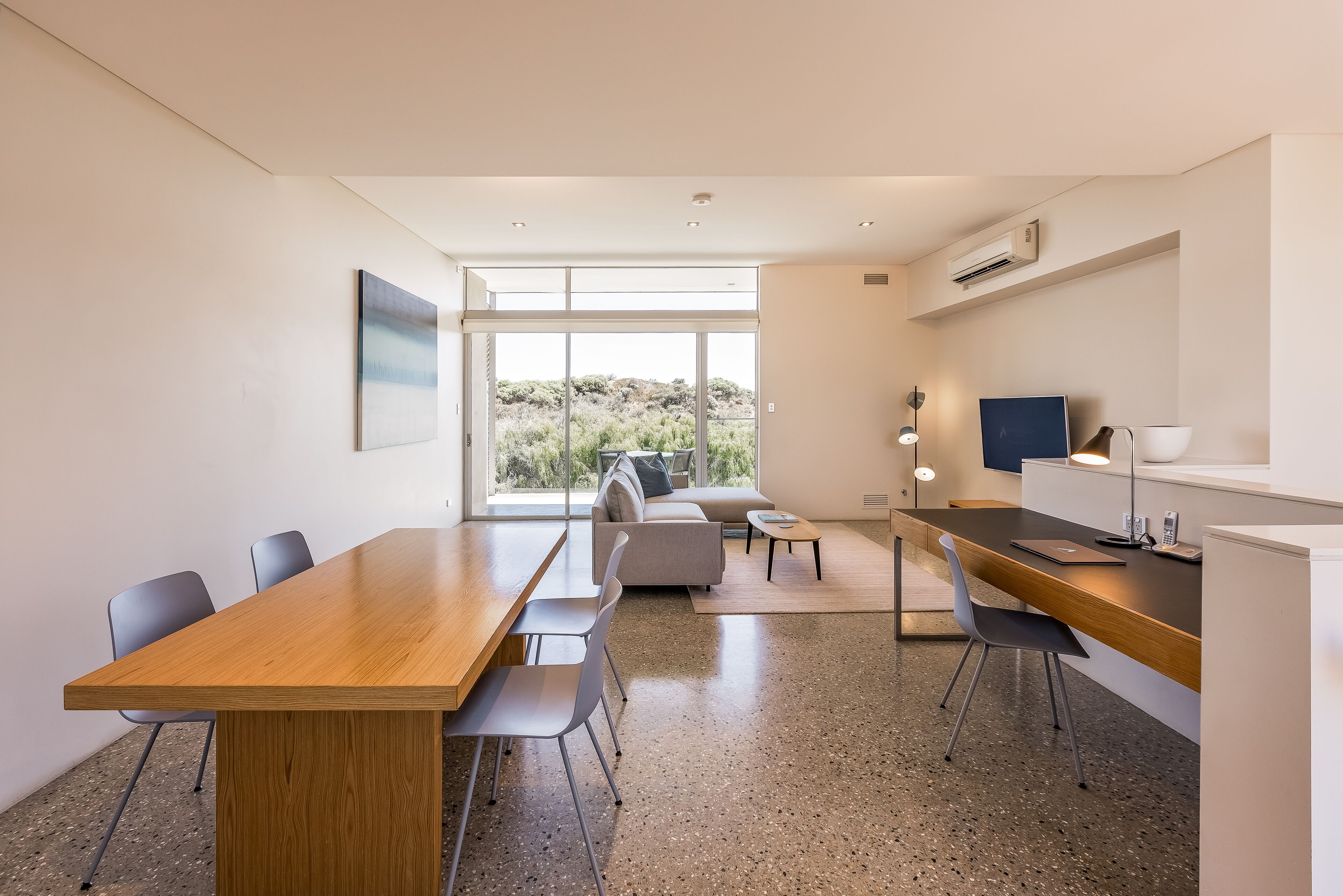 A modern, well-lit room with a wooden dining table, chairs, a sofa, and a desk with a computer.