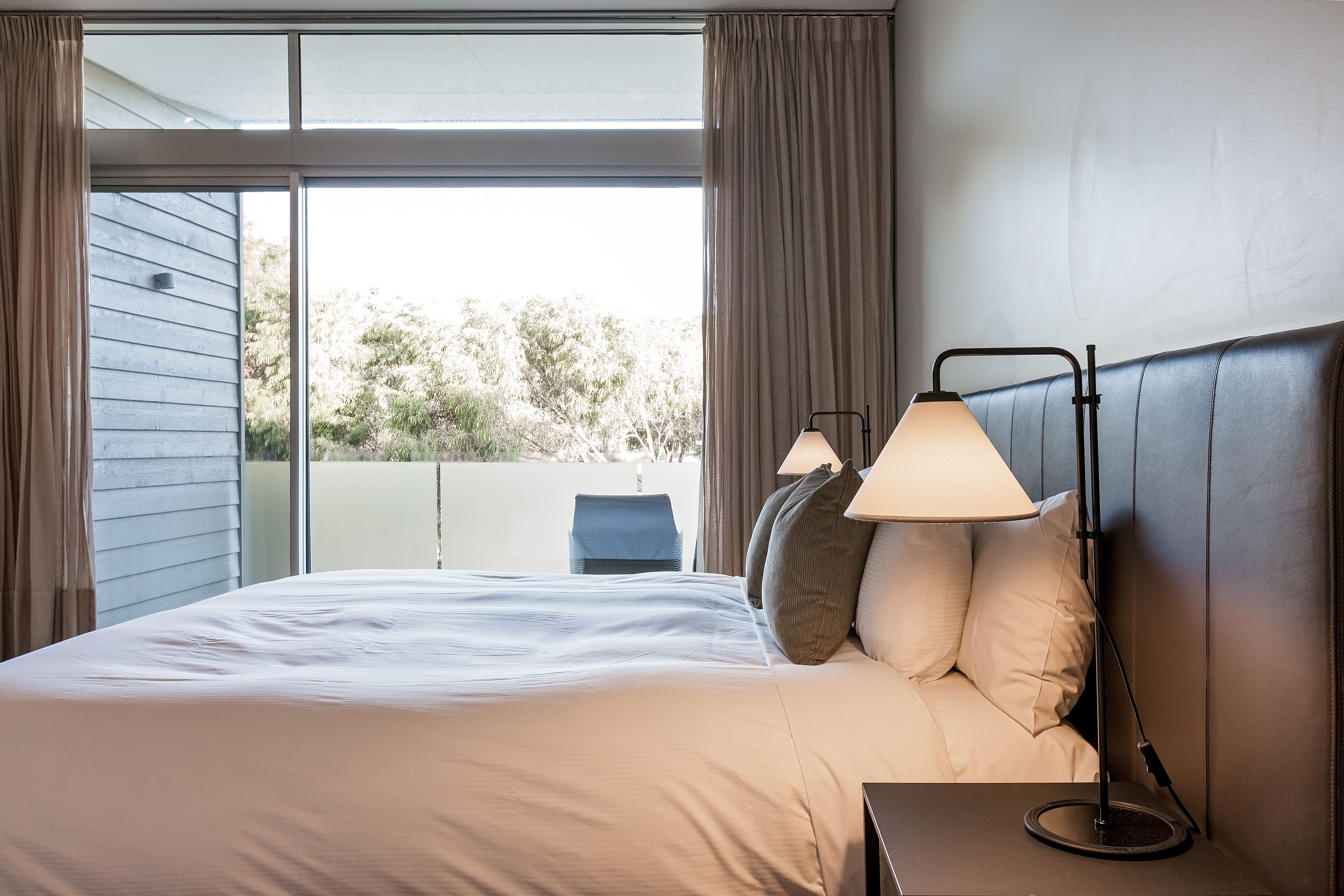 Uma cama bem arrumada com cabeceira, duas luminárias de cabeceira e vista para o exterior por uma grande janela.