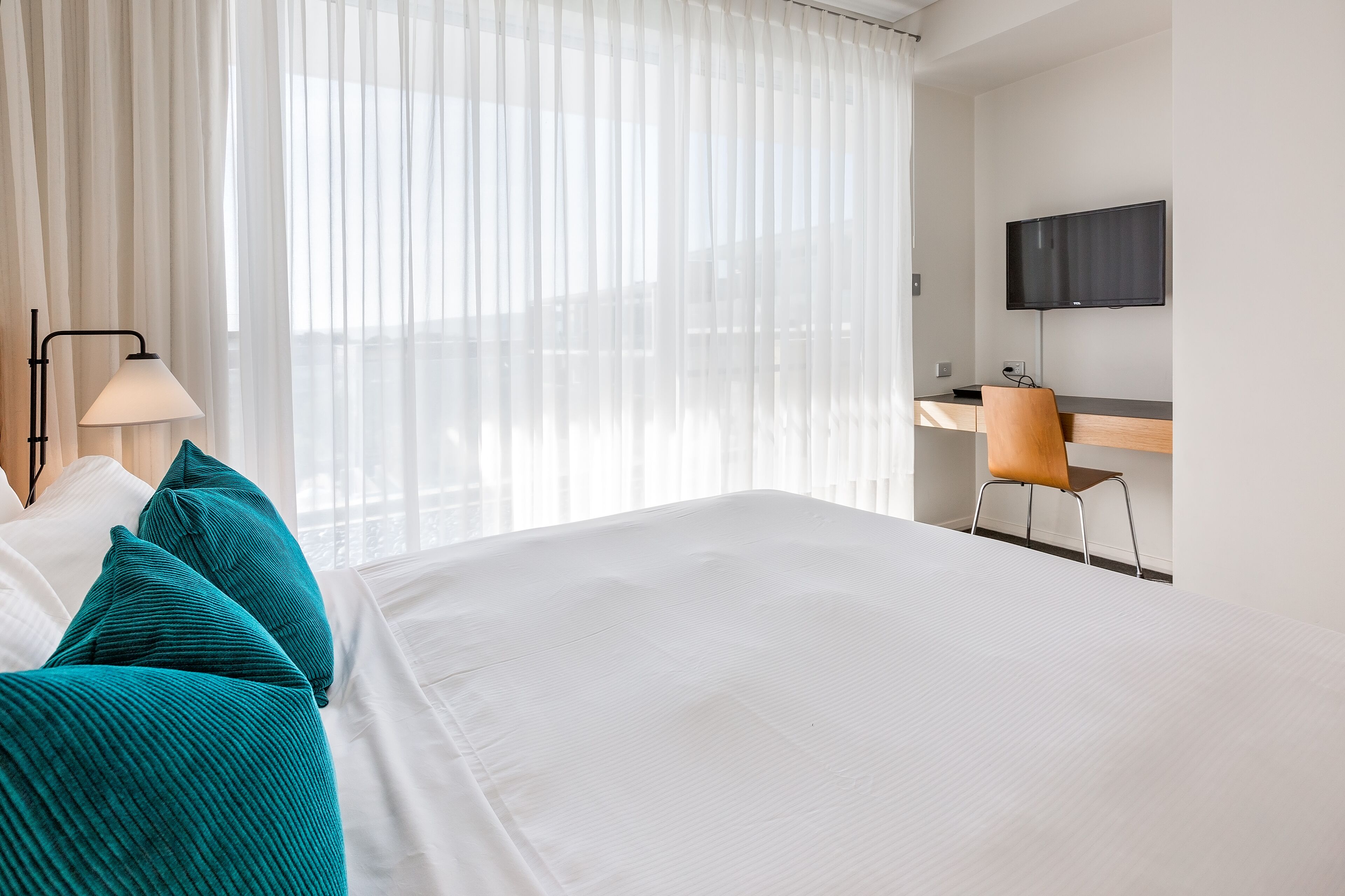 A hotel room with a large bed, a desk with a chair, a TV mounted on the wall, and sheer curtains.