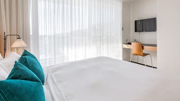 A hotel room with a large bed, a desk with a chair, a TV mounted on the wall, and sheer curtains.