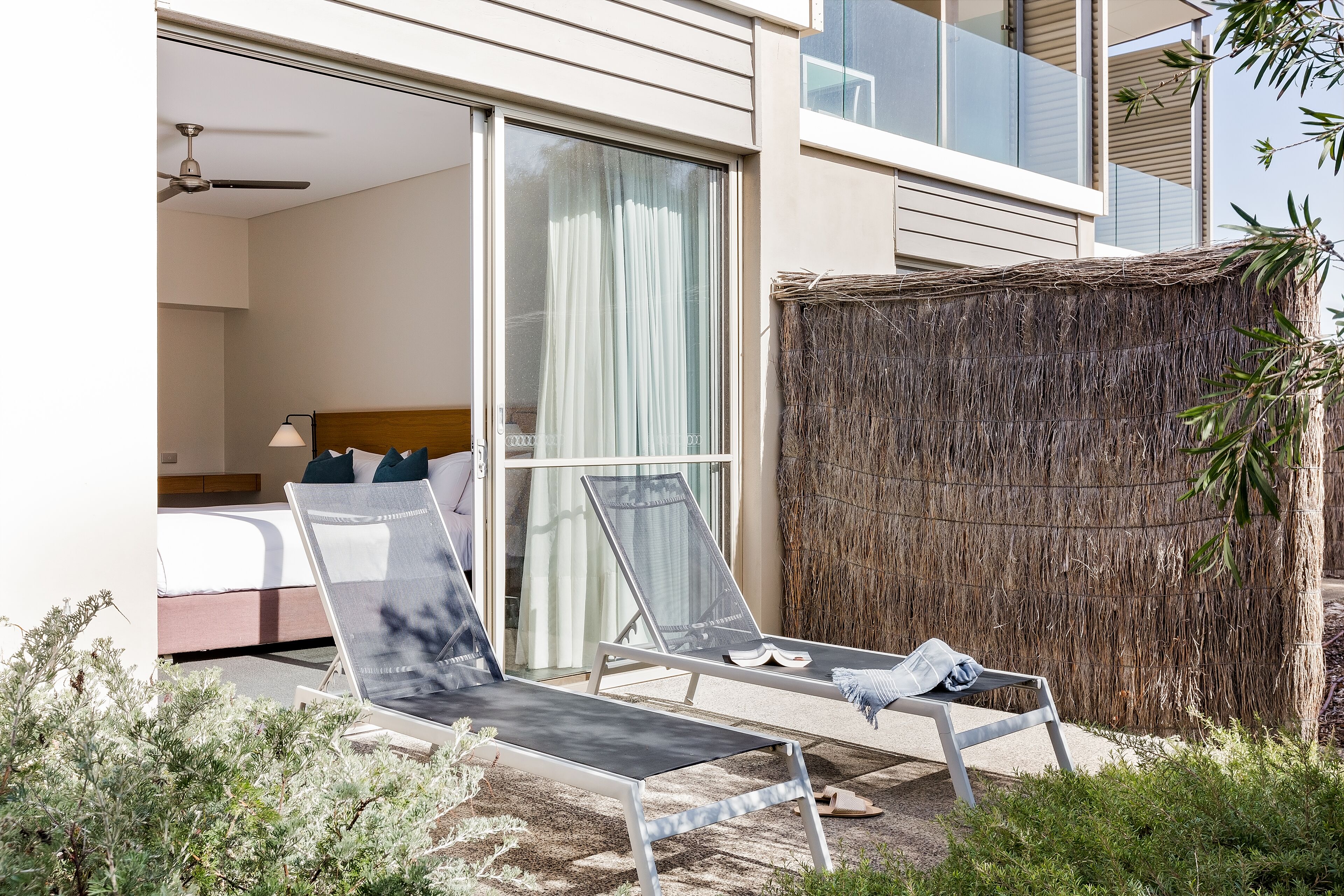 A modern hotel room with a balcony, a bed with pillows, a ceiling fan, and a large window with curtains.