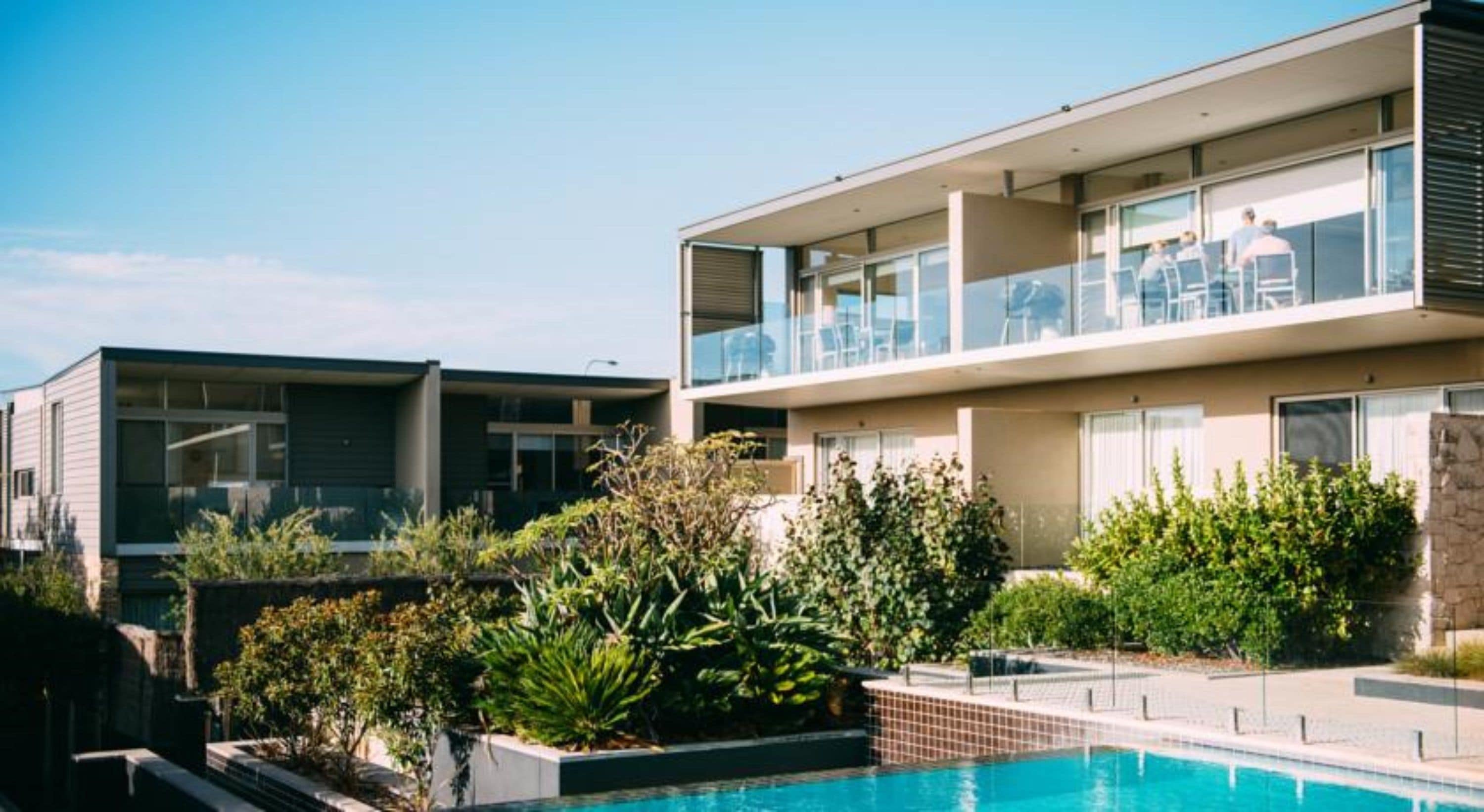 A modern two-story residential building with a swimming pool, surrounded by lush greenery and a clear sky.