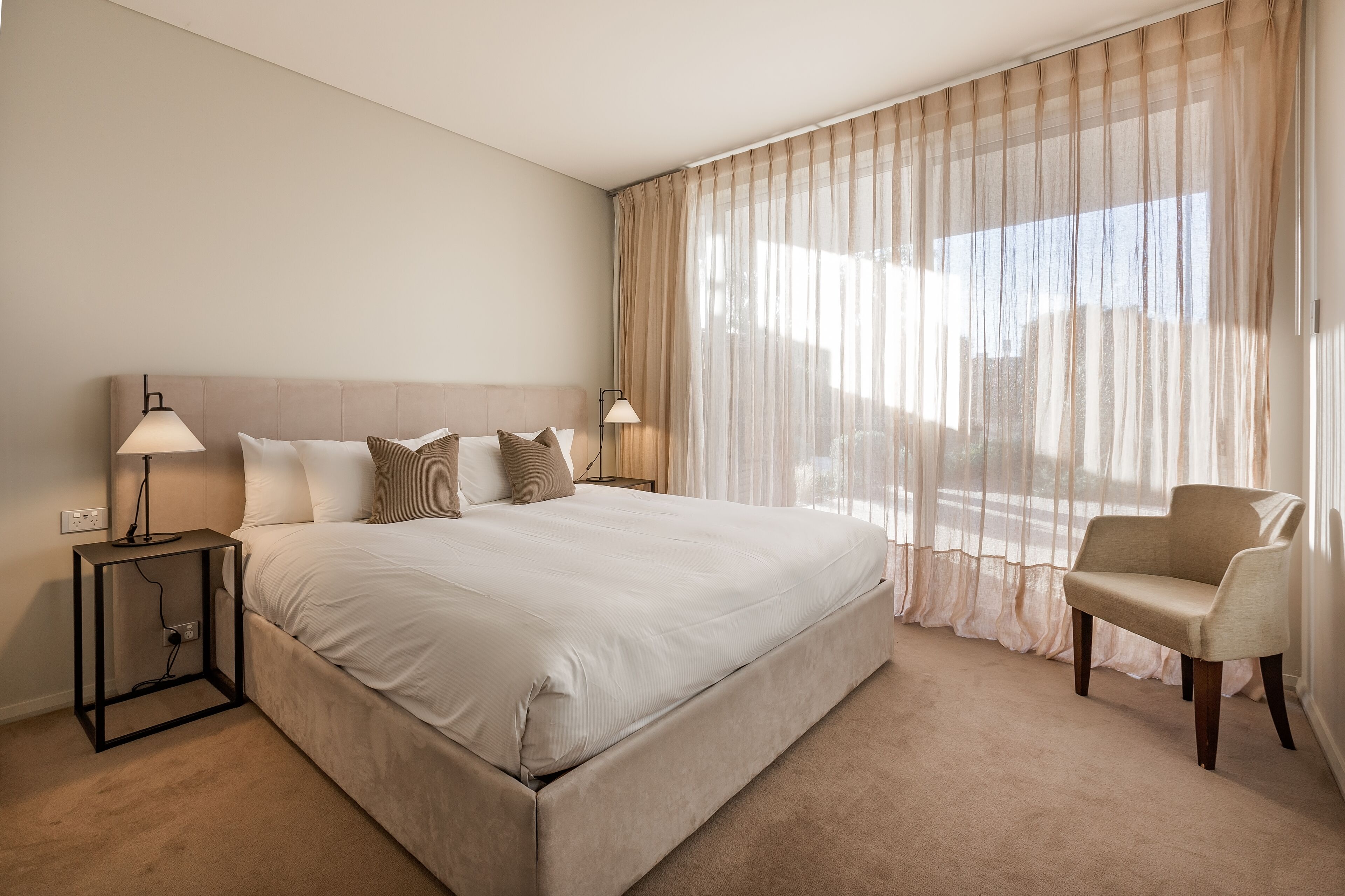 Ein Hotelzimmer mit einem großen Bett, zwei Nachttischlampen, einem Sessel und durchsichtigen Vorhängen.