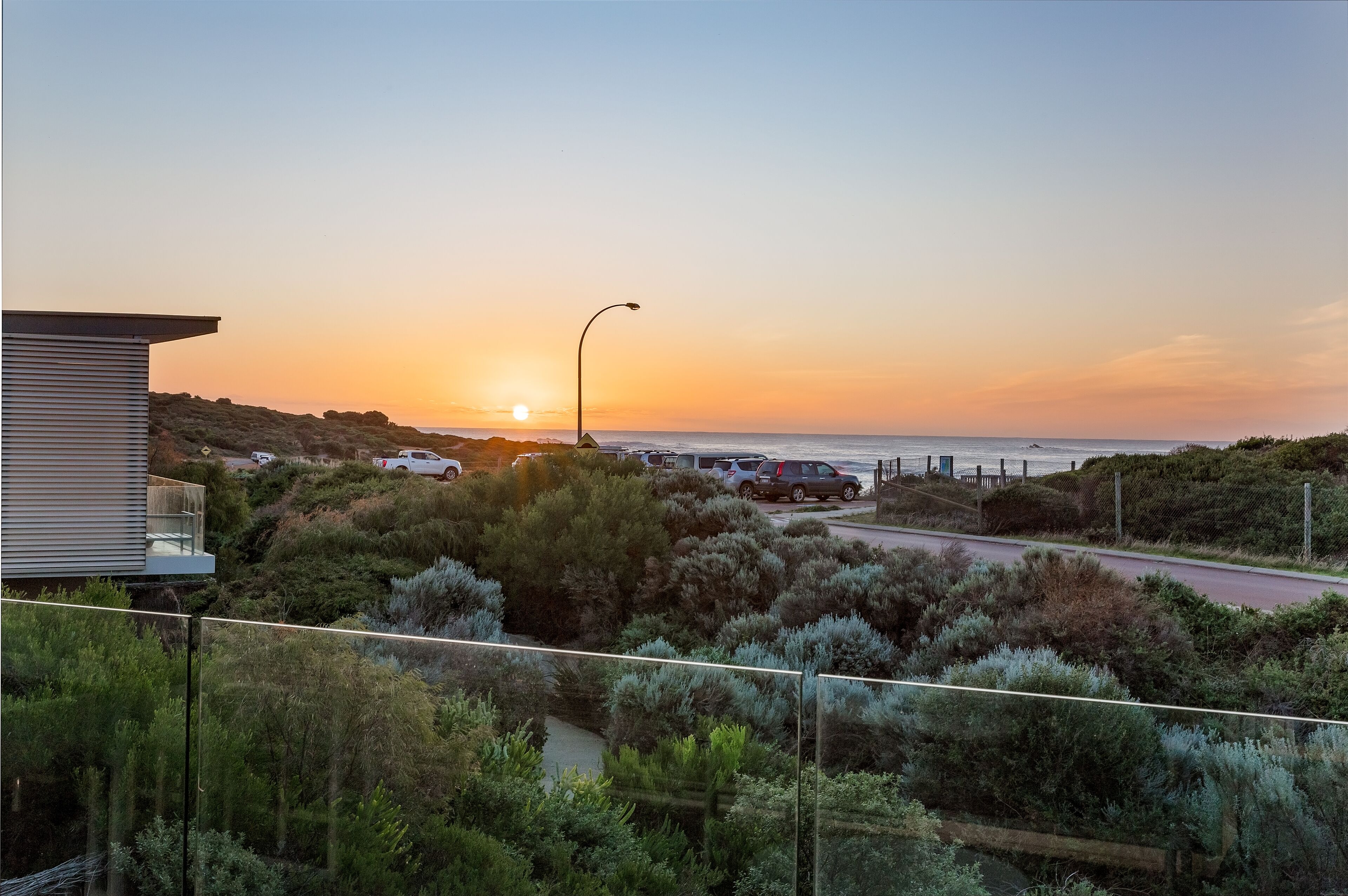 Paesaggio costiero con tramonto, una strada, auto parcheggiate e un edificio con balcone che si affaccia sul mare.