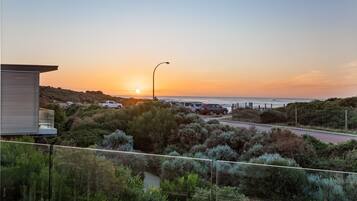 Paesaggio costiero con tramonto, una strada, auto parcheggiate e un edificio con balcone che si affaccia sul mare.