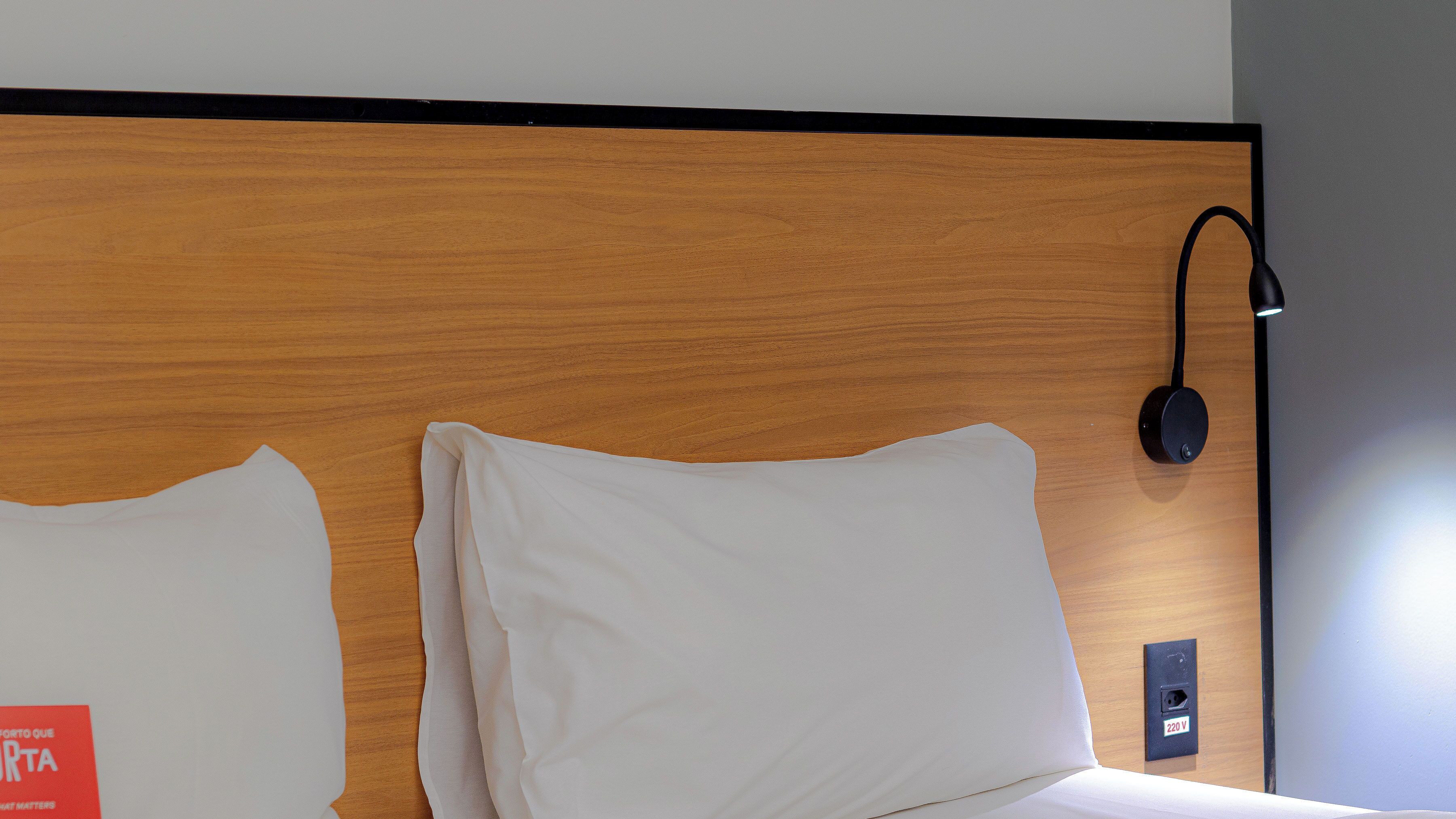 Habitación de hotel con cabecera de madera, almohadas blancas, lámpara negra fijada a la pared y un enchufe con la etiqueta 220V.