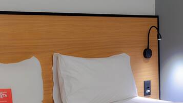 Habitación de hotel con cabecera de madera, almohadas blancas, lámpara negra fijada a la pared y un enchufe con la etiqueta 220V.