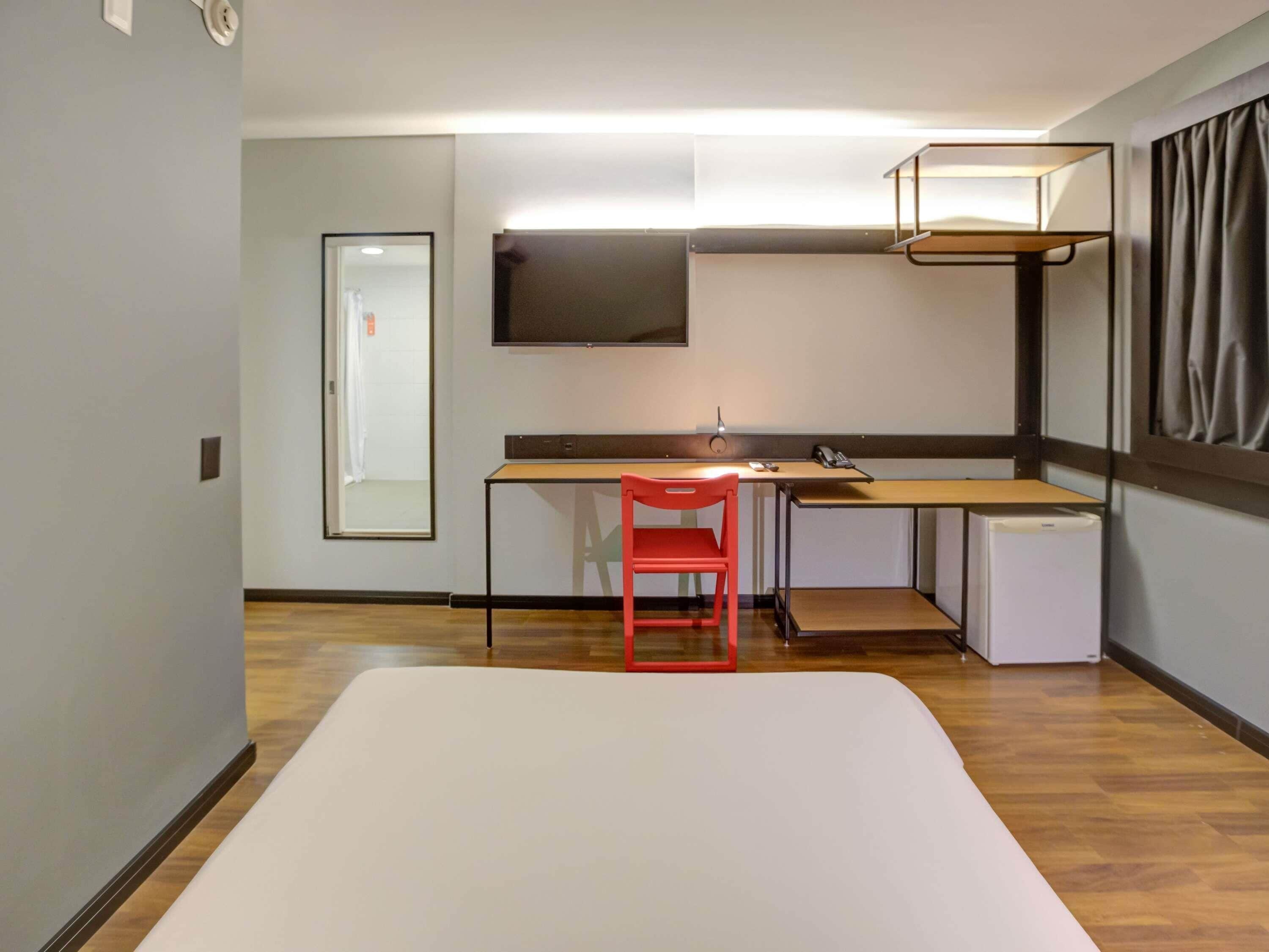 A compact hotel room with a bunk bed, a desk with a chair, a small refrigerator, a flat-screen TV, and a window with curtains.