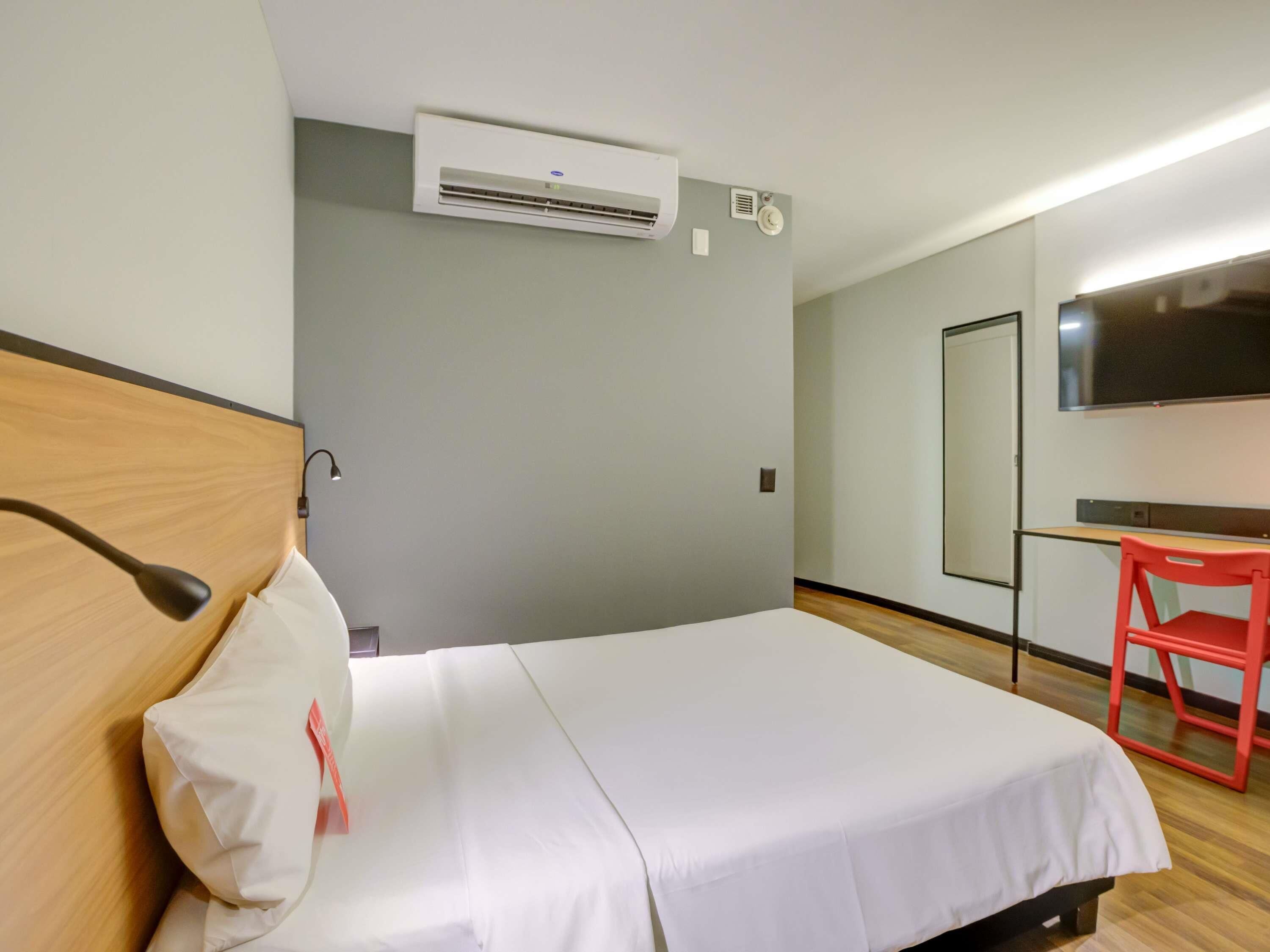 A hotel room with a bed, a desk, a chair, an air conditioning unit, and a television on the wall.