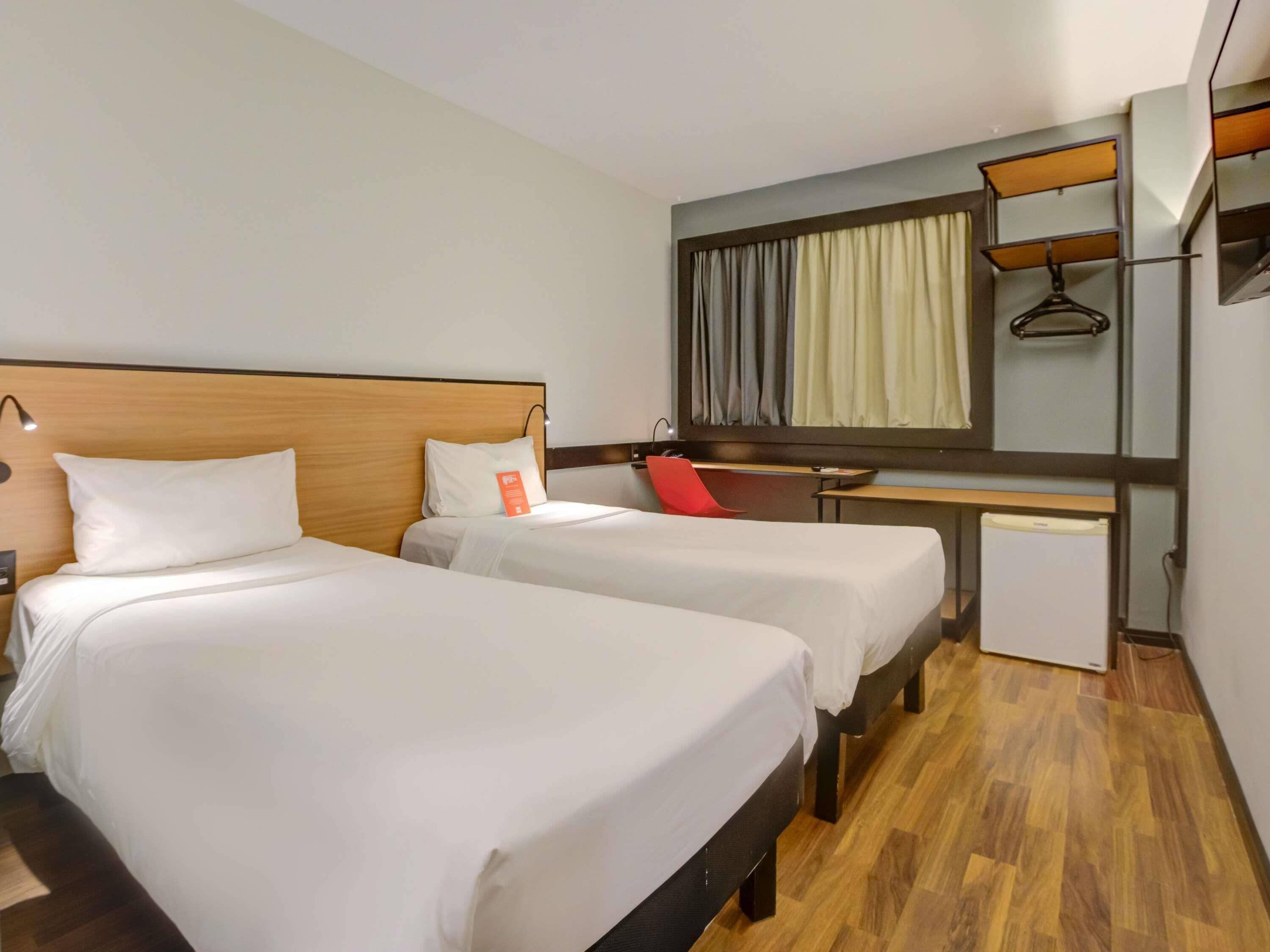 Habitación de hotel con dos camas, un escritorio, una silla, un refrigerador pequeño y un espejo.