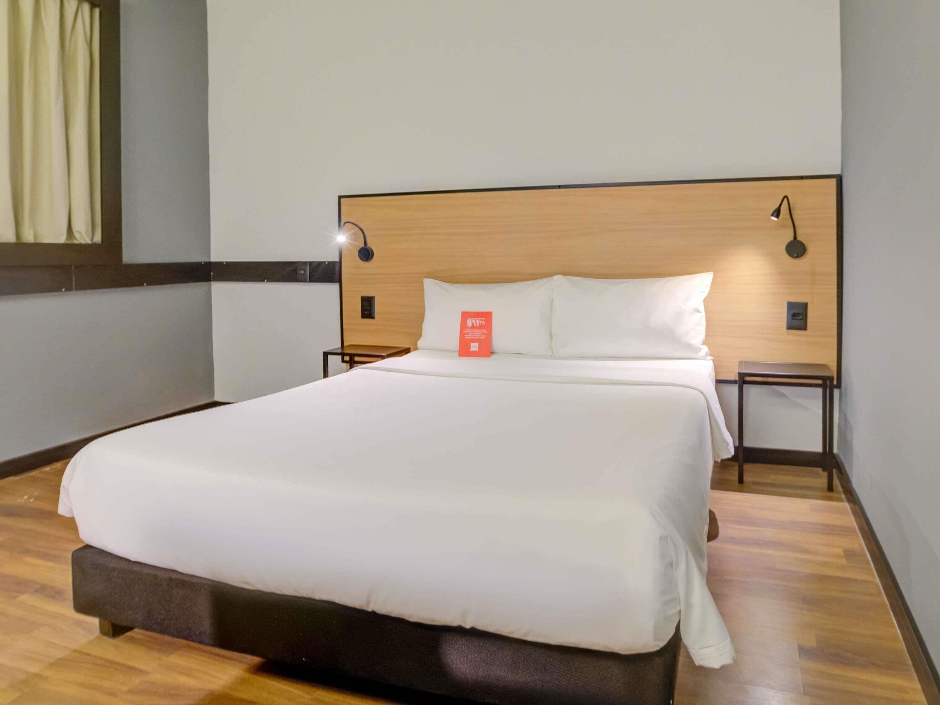 A neatly made bed in a hotel room with a wooden headboard, two bedside tables, and a wall-mounted lamp.