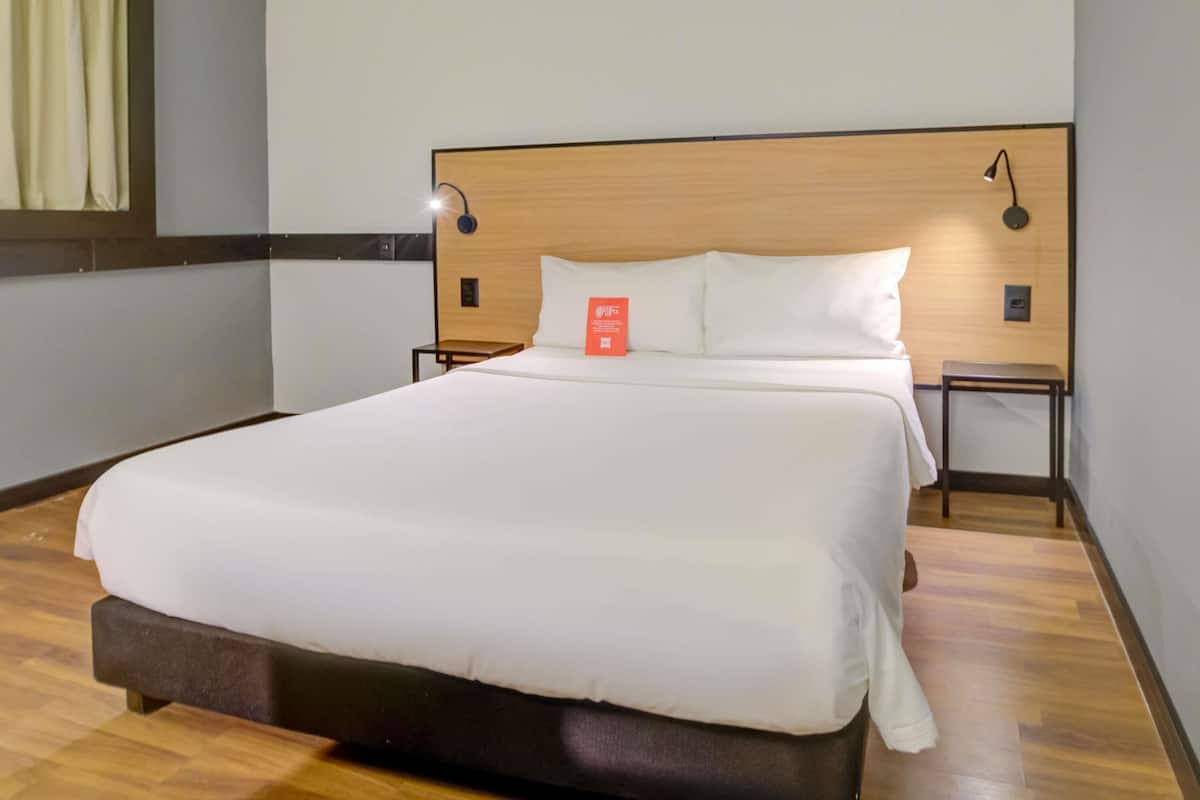 A neatly made bed in a hotel room with a wooden headboard, two bedside tables, and a wall-mounted lamp.