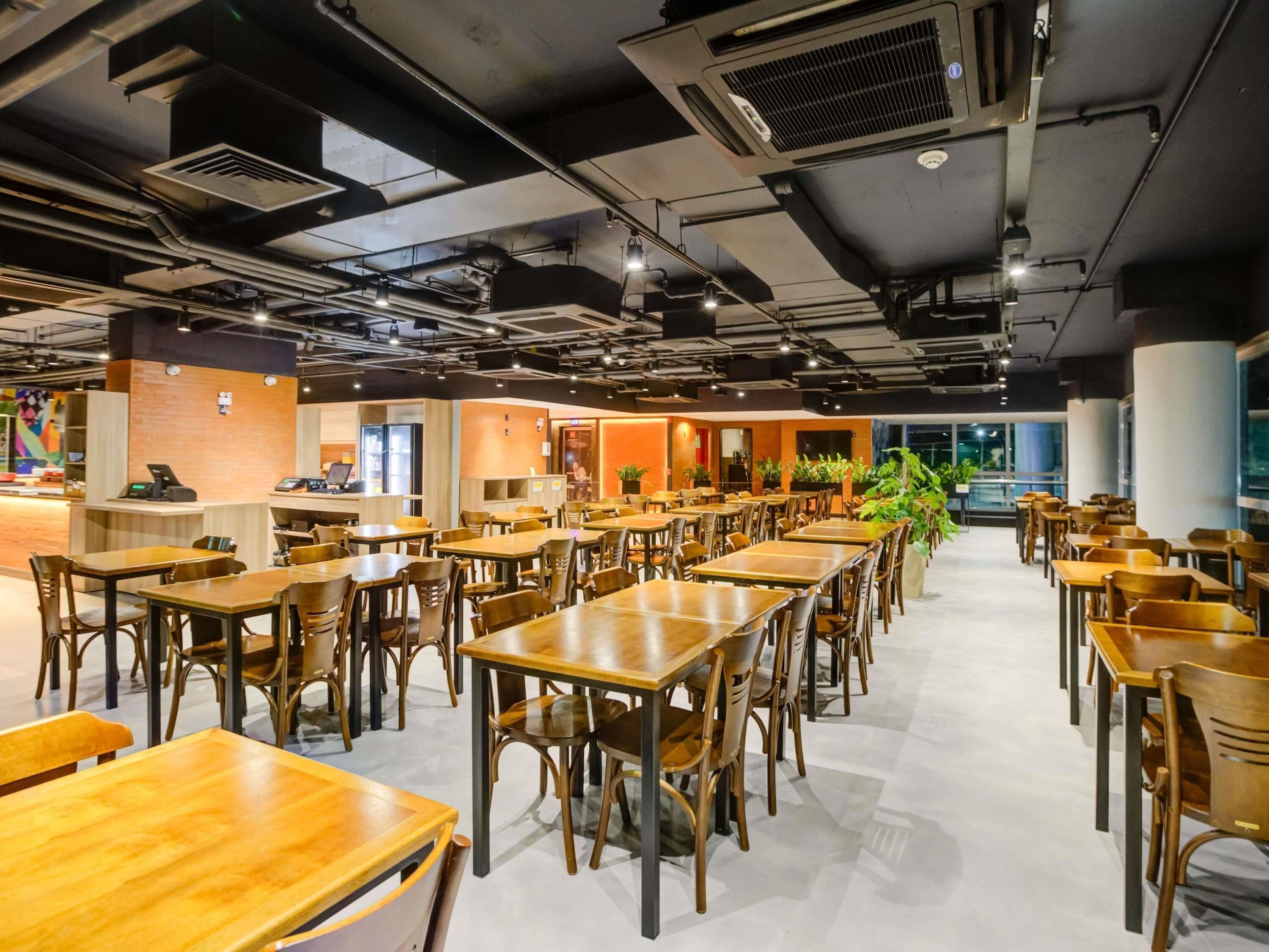 Un vaste espace de restauration intérieur, doté de nombreuses tables et chaises en bois, d’un comptoir avec une caisse enregistreuse et de plantes en pot.