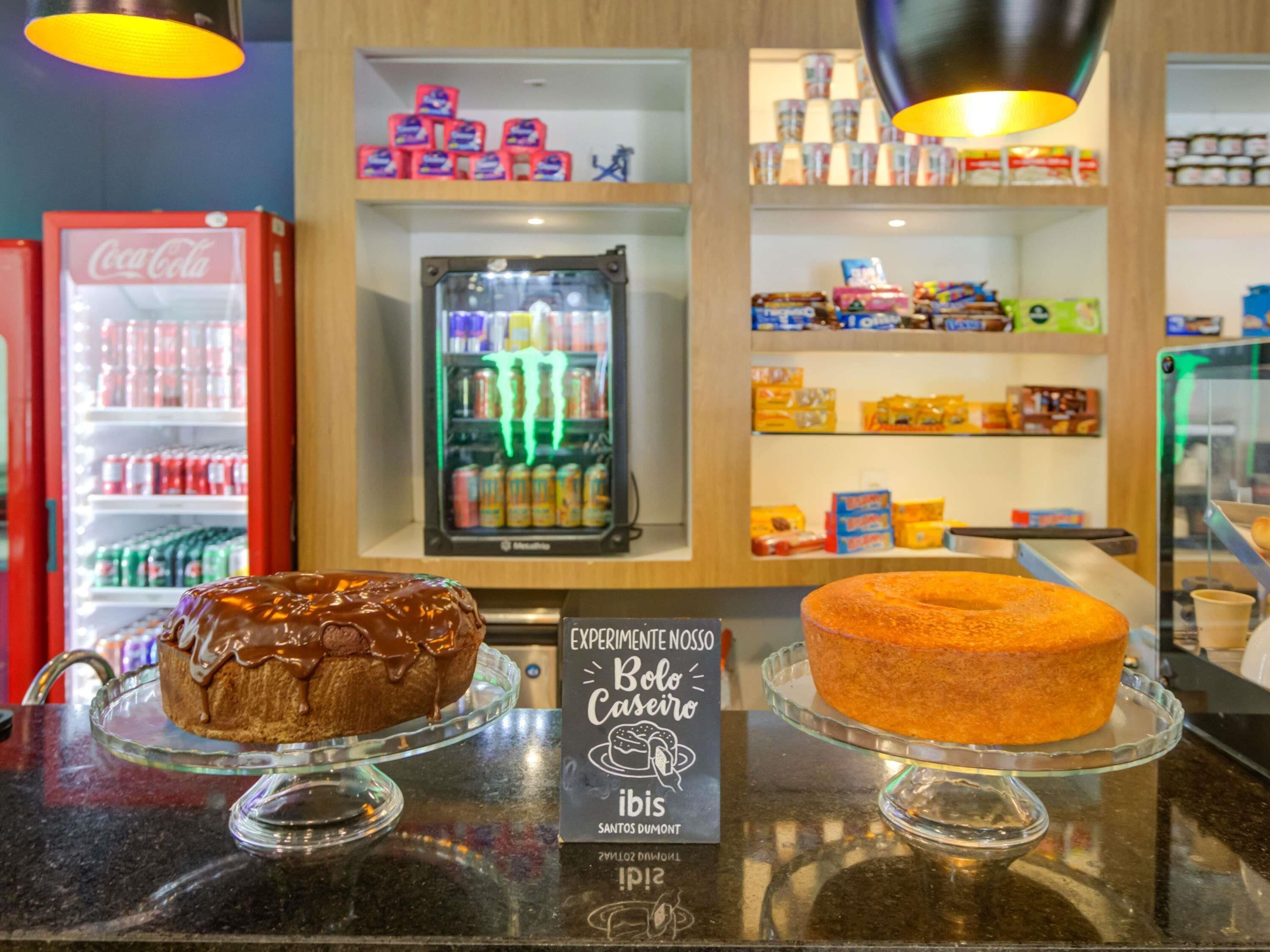 A bakery display with two cakes on glass stands, a Coca-Cola refrigerator, and shelves stocked with various products.